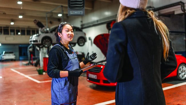 Female mechanic delivering repaired car to customer in workshop Professional female mechanic explaining car repair details to customer, holding clipboard in auto repair shop
